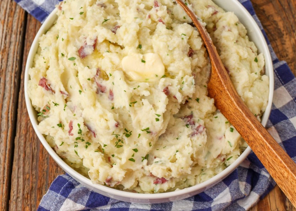 mashed red potatoes in white bowl with wooden spoon and blue napkin