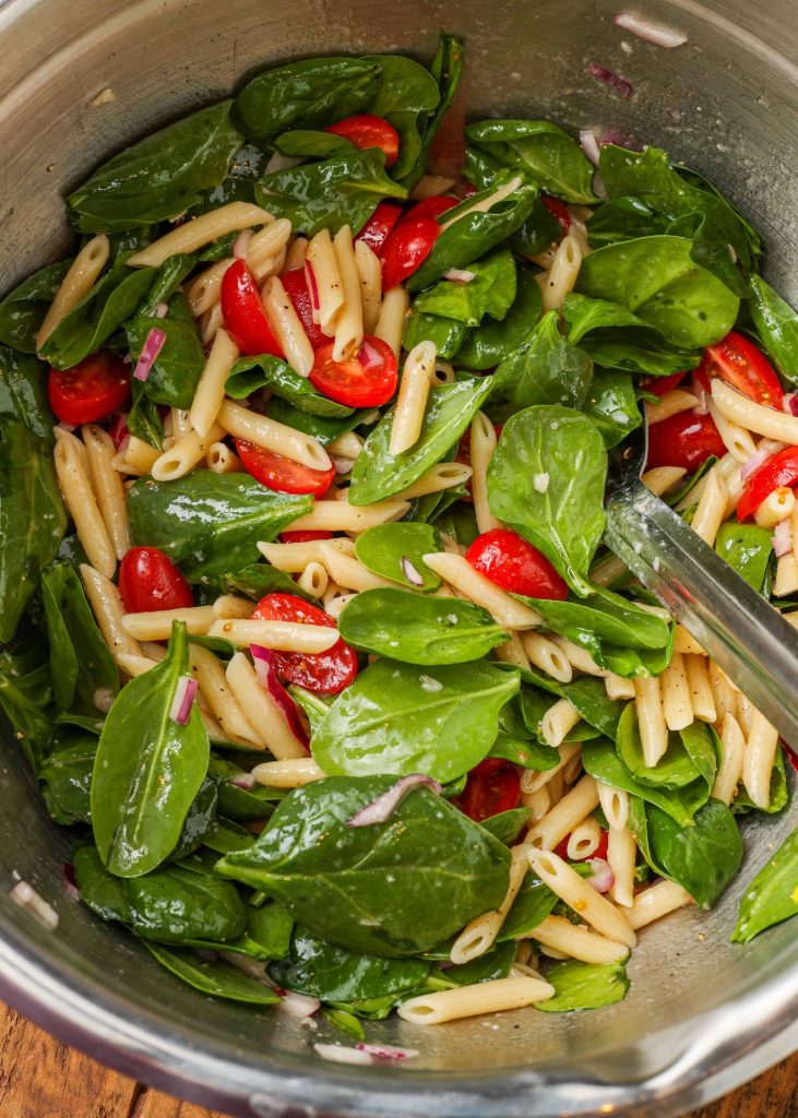 spinach salad with pasta and tomatoes in metal bowl with serving spoon