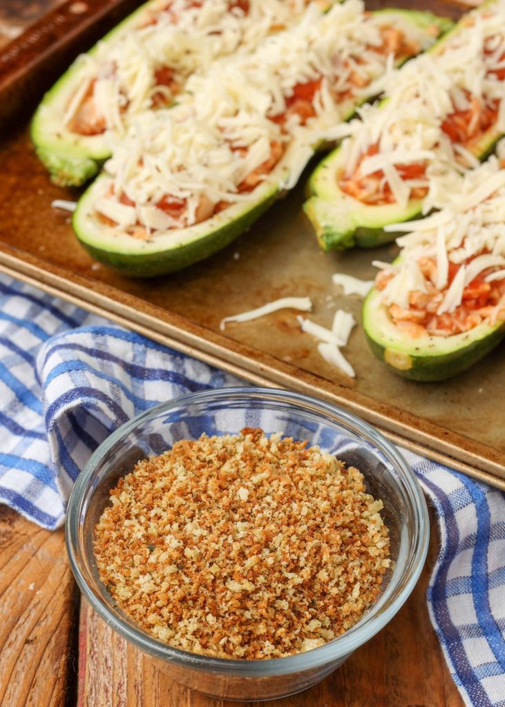 a glass bowl of toasted bread crumbs sits next to a sheet pan with zucchini boats filled and ready for topping