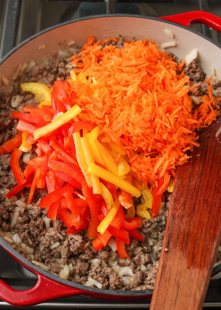 matchstick chopped multicolor bell peppers and carrots have been placed atop cooked ground beef and onions in this red skillet with a white interior