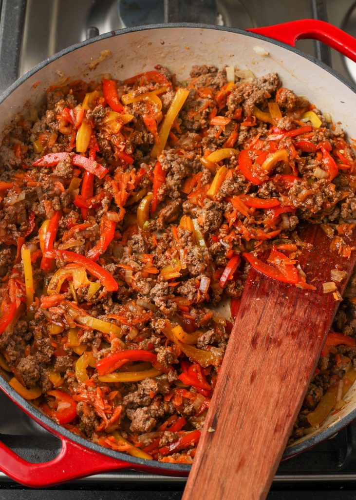 All of the ground beef and vegetables have been cooked in a red skillet with a wooden spatula sticking out of it