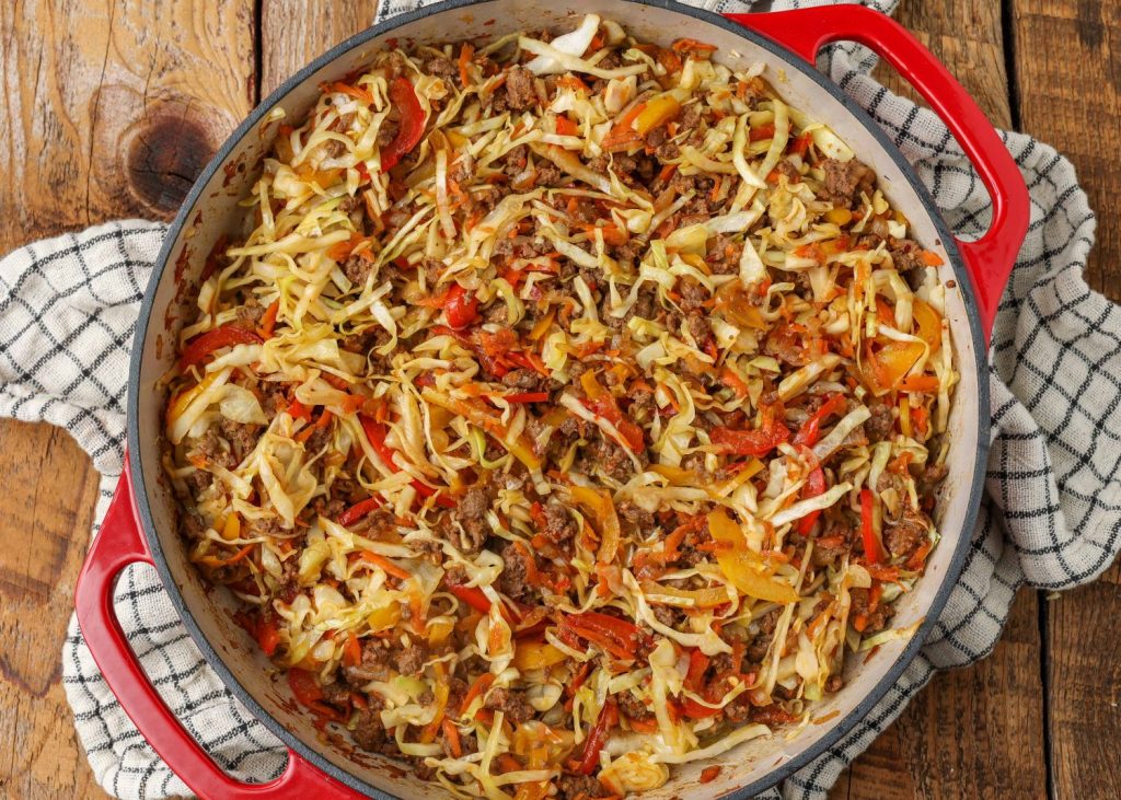 a top down photo of a red skillet with a white interior, full of ground beef and cabbage with match stick carrots and multicolor bell peppers