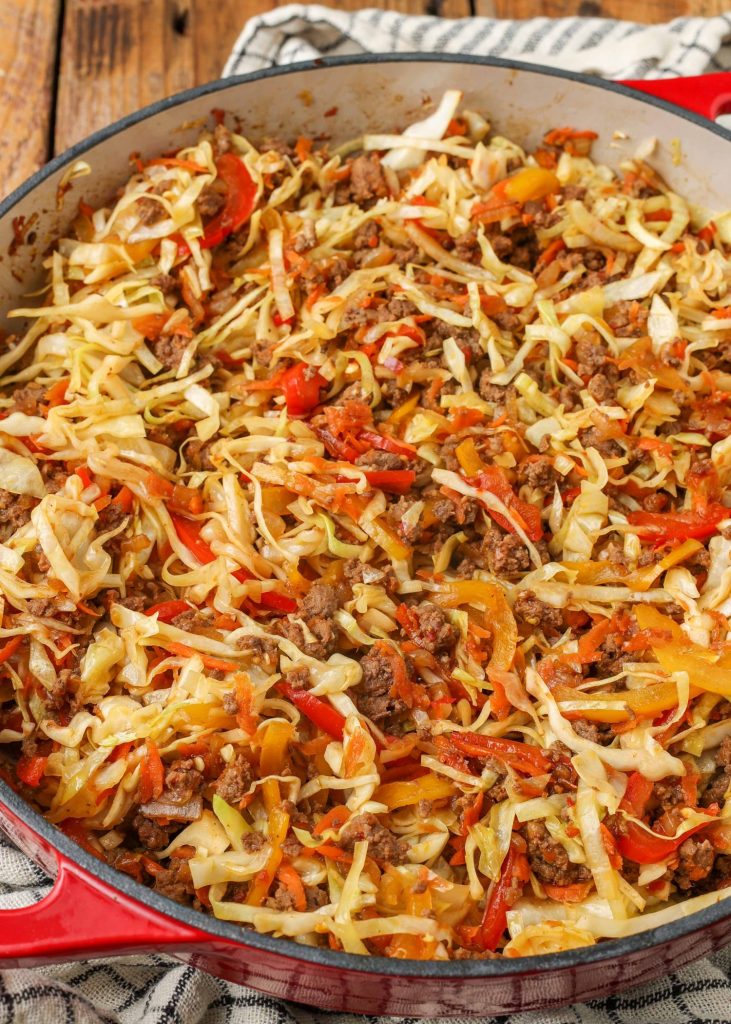 a close up photo of ground beef, cabbage, carrots, and bell peppers with onions and garlic in the skillet