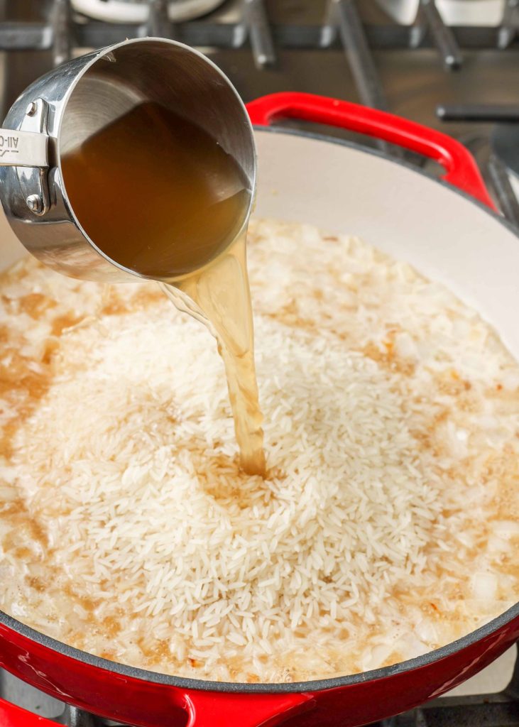 chicken broth is poured out of a metal measuring cup onto uncooked rice grains in a red skillet