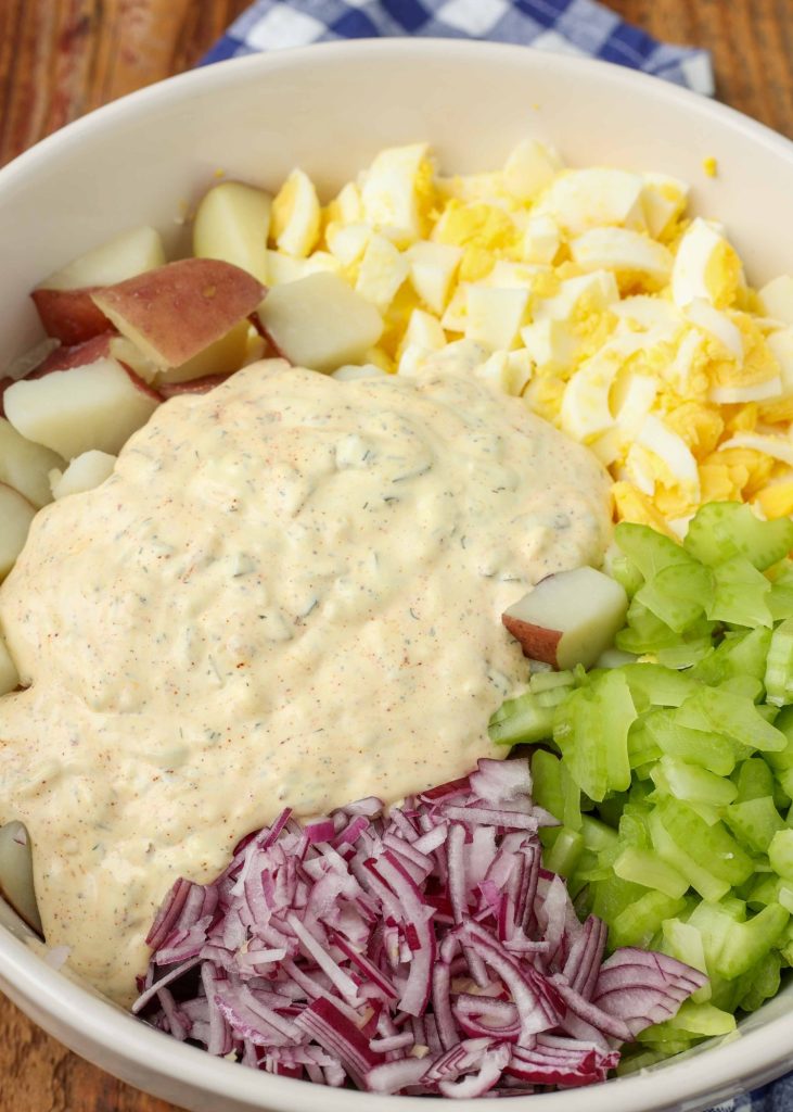A bowl with all of the ingredients ready to combine to make the red skin potato salad with bright purple onions, green celery, and yellow egg yolks