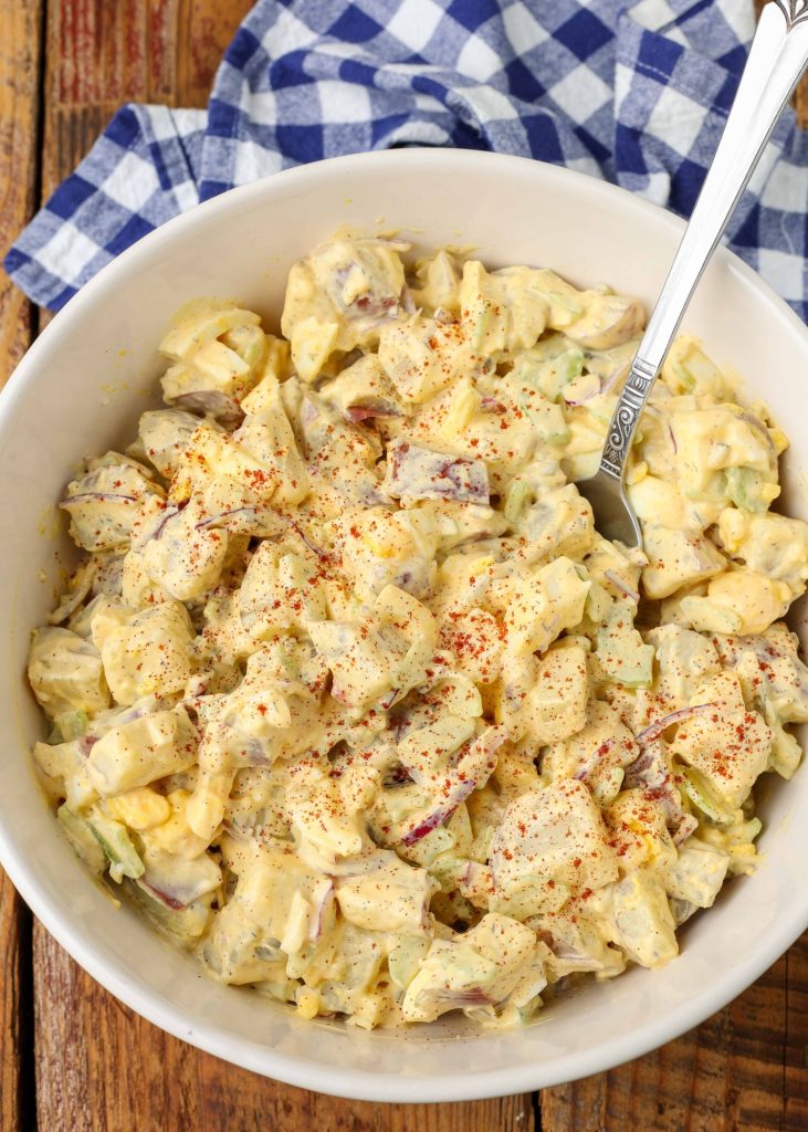 A top down photo of a bowl of potato salad that has been sprinkled with paprika and is ready to eat with the metal spoon in it