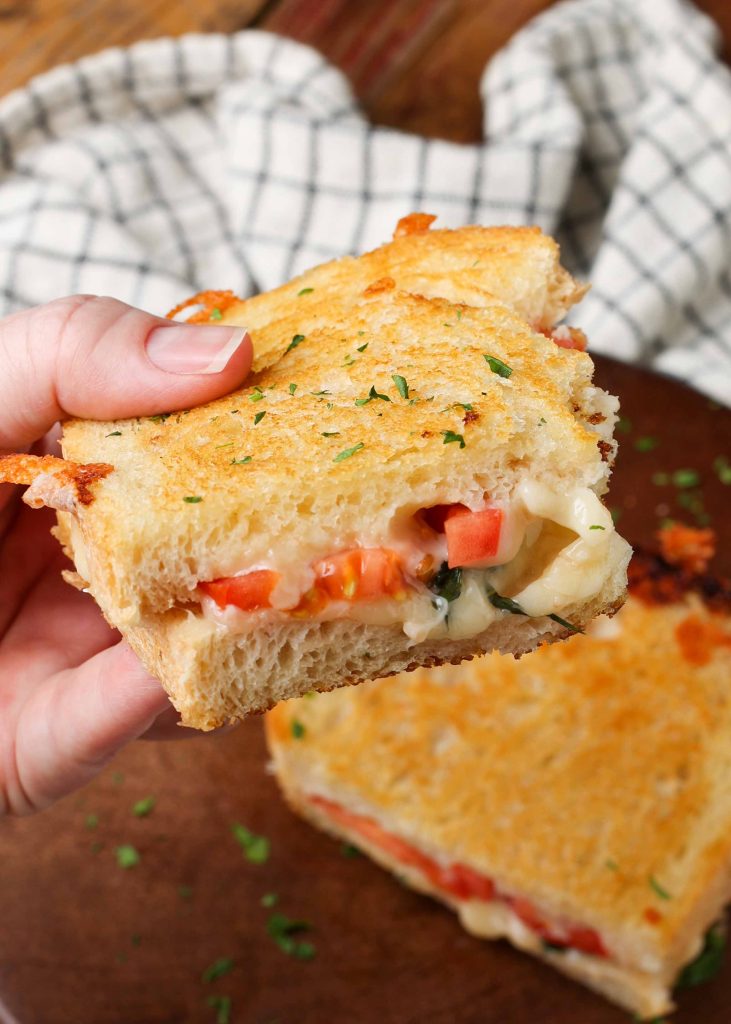 a hand holds half of the grilled cheese sandwich with tomatoes above the table with a bite missing