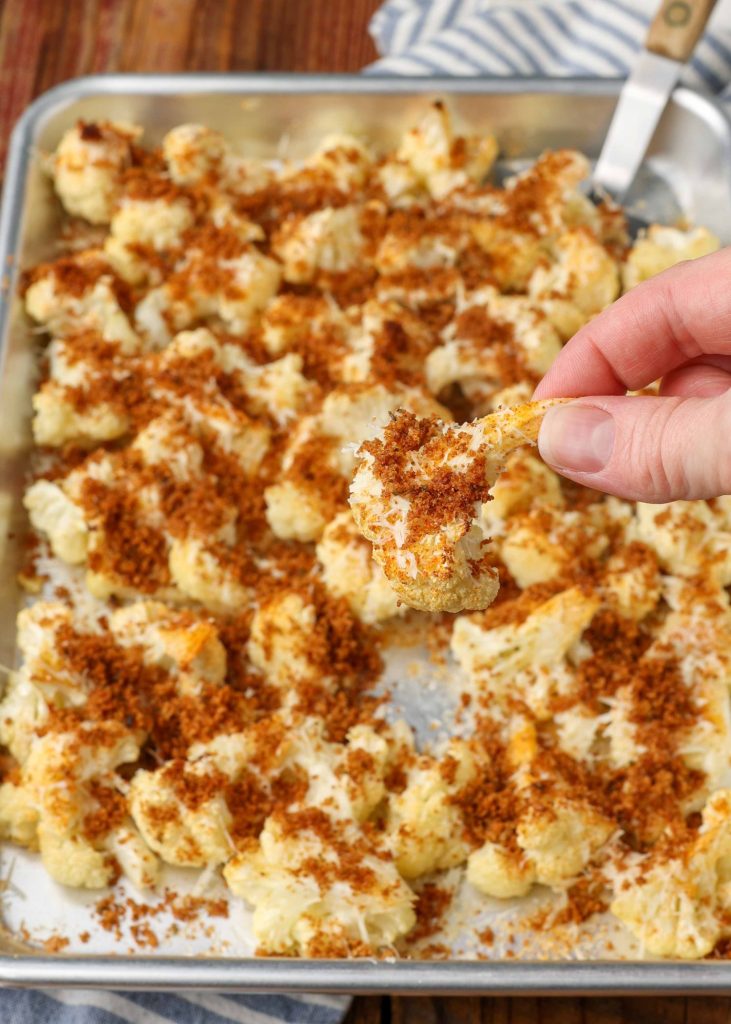 fingers have lifted a floret of cauliflower off of the metal sheet pan in the background. it is sprinkled with toasted bread crumbs and savory nutty parmesan.
