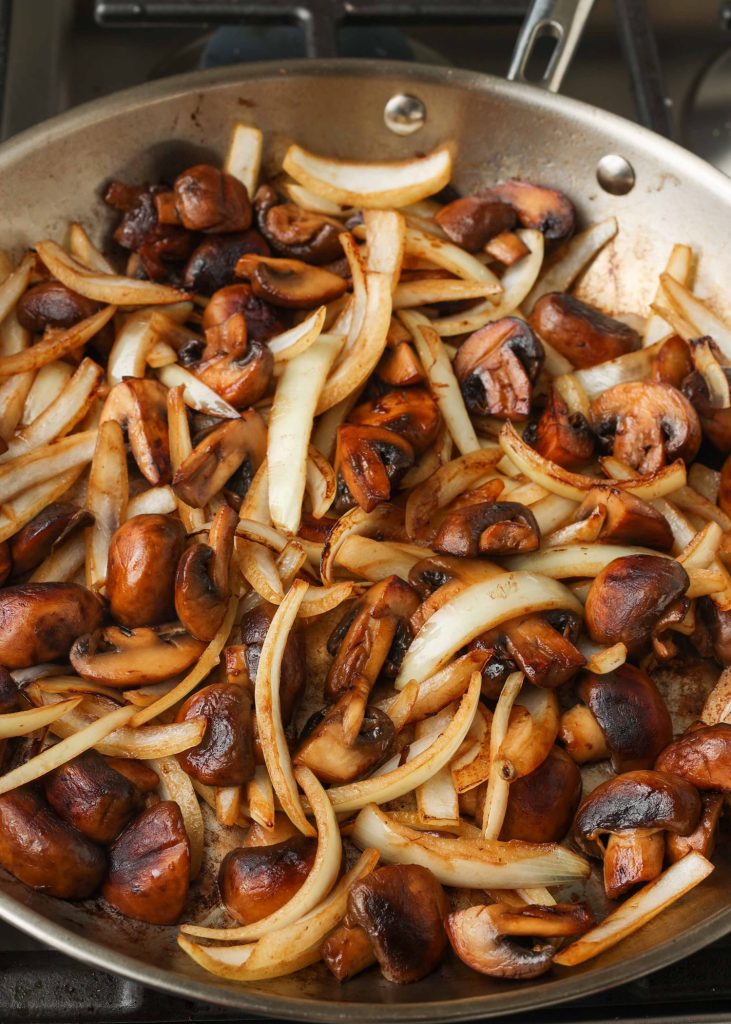 raw onions have been added to the skillet of mushrooms that have cooked down and are ready to be seared