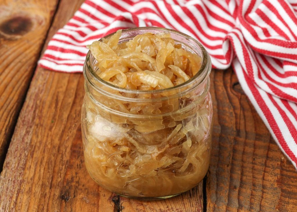 a close up photo of caramelized onions in a small jar on a wooden tabletop with a red and white striped napkin in the background