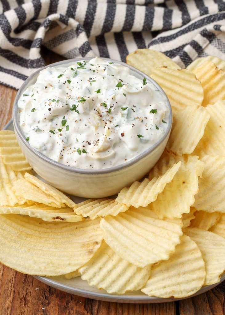 a small glass bowl of artichoke jalapeno dip beside a pile of ruffled potato chips on a plate