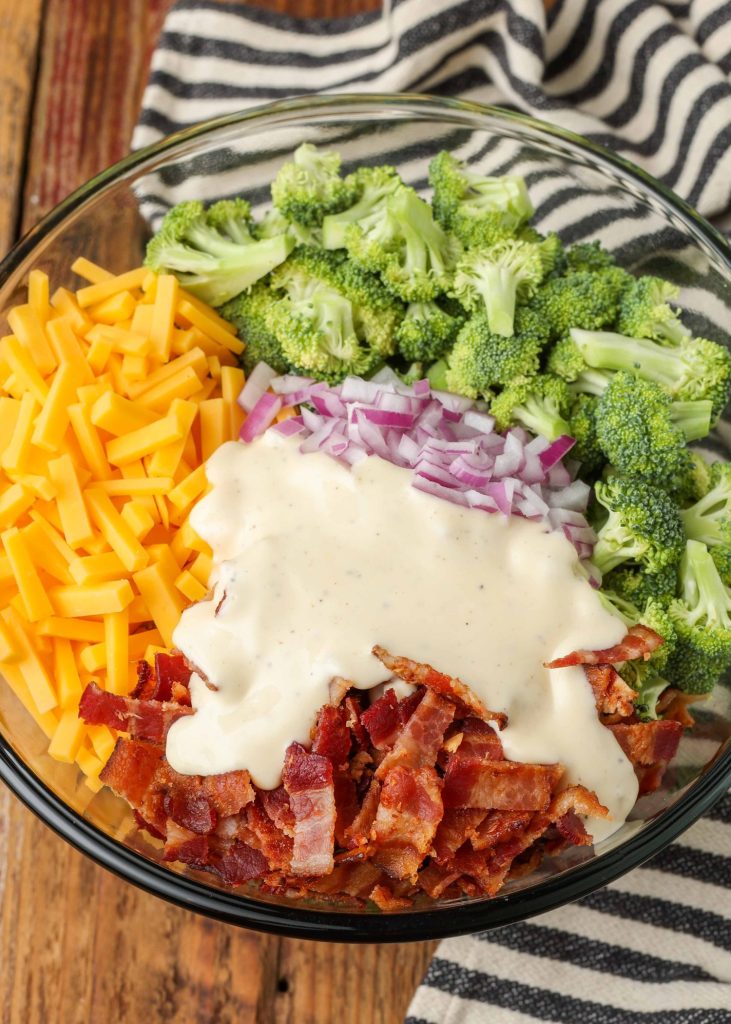 Overhead shot of broccoli, onions, bacon, and cheese in a tall glass bowl, all chopped into bite-size pieces, topped with creamy dressing