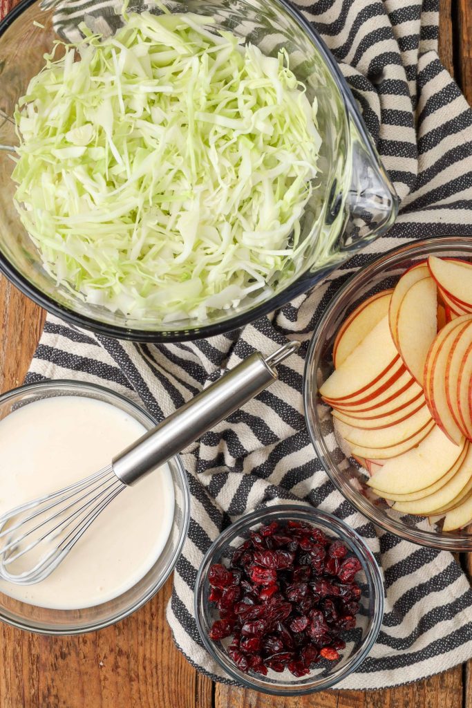 Overhead shot of sliced apples, cabbage, cranberries, and savory dressing, each served in a glass bowl