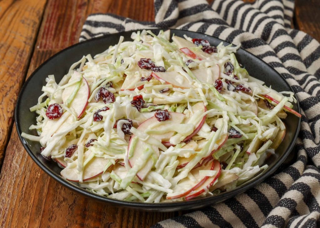 Overhead horizontal shot of cranberry apple coleslaw, served in a black bowl with a striped white and gray towel