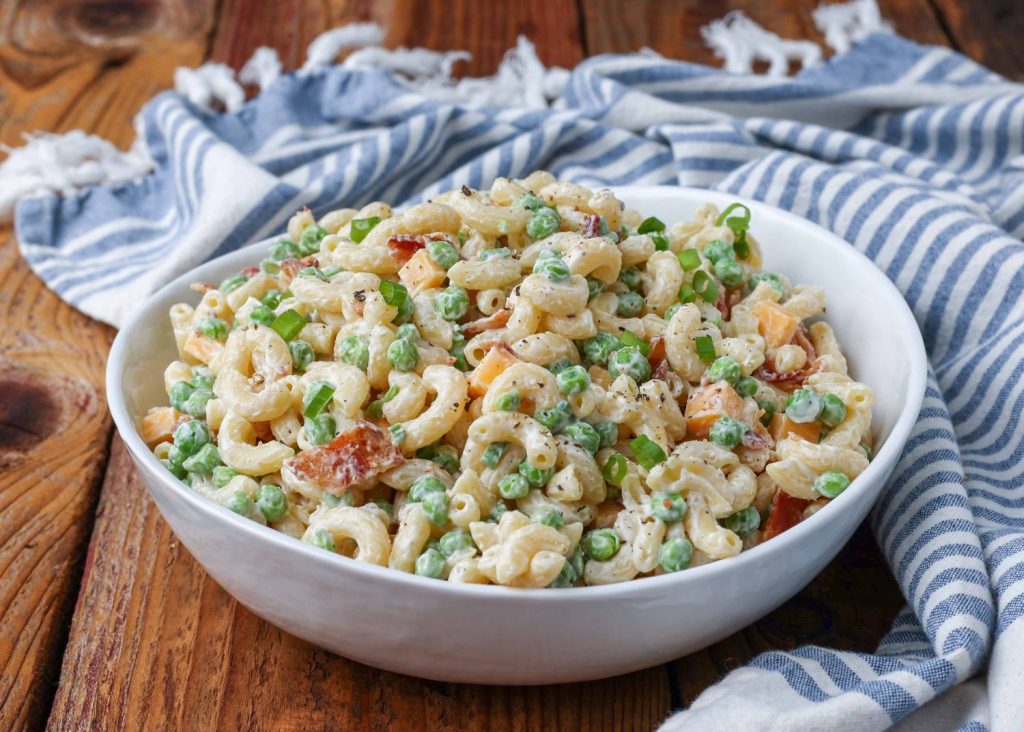 Horizontal shot of creamy pea pasta salad with bacon and cheese in a white bowl with a striped blue and white towel
