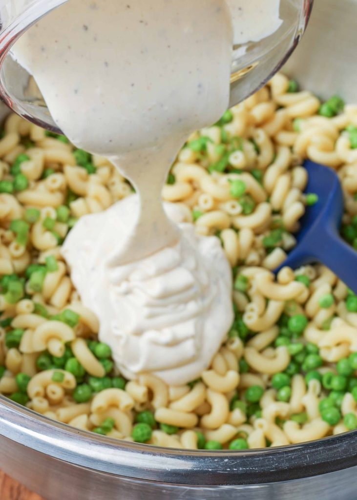 Pasta and peas in a stainless steel bowl with a blue rubber spatula; creamy mayo is being poured into the bowl
