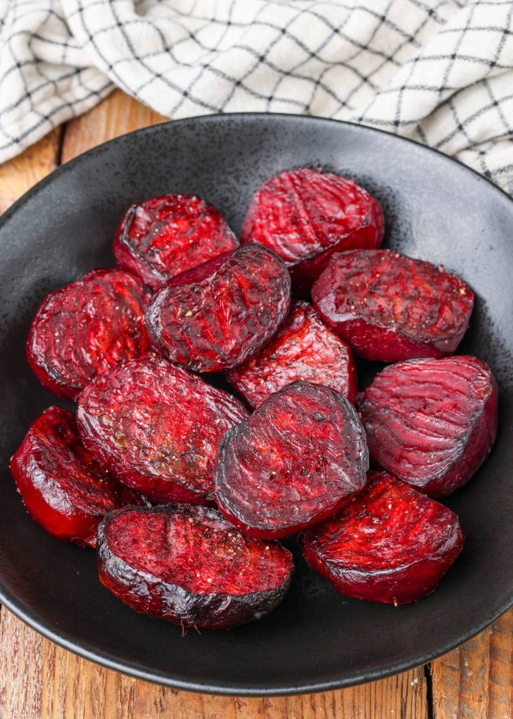 a black plate full of sliced roasted beets on a wooden tabletop with a checkered black and white napkin visible in the background