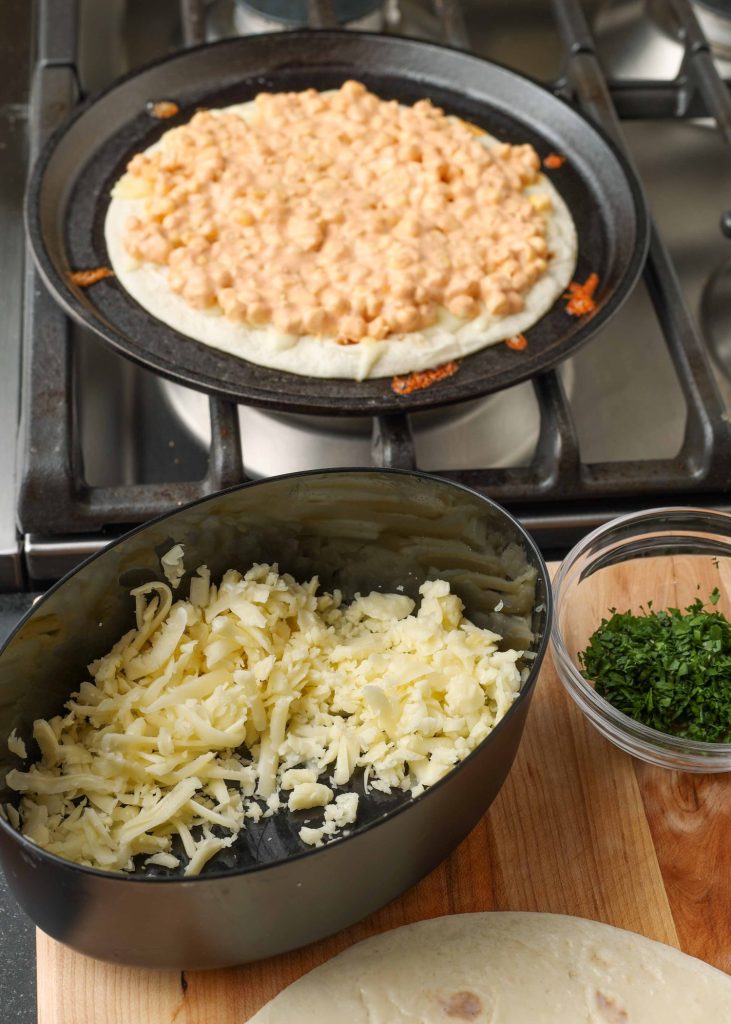 Overhead shot of tortilla topped with corn mixture cooking in a skillet; next to the stove is a tub filled with shredded cheese and a glass bowl filled with cilantro