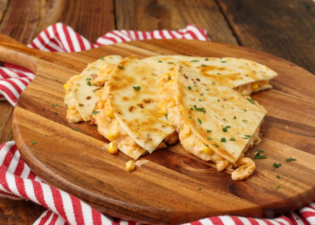 Wide shot of a cheesy corn quesadilla topped with cilantro, cut into quarter wedges and served on a round wooden cutting board on a striped red and white towel