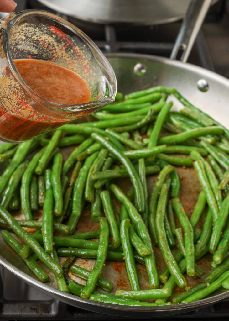 Overhead shot of spicy red peanut sauce being poured into a stainless steel skillet filled with green beans