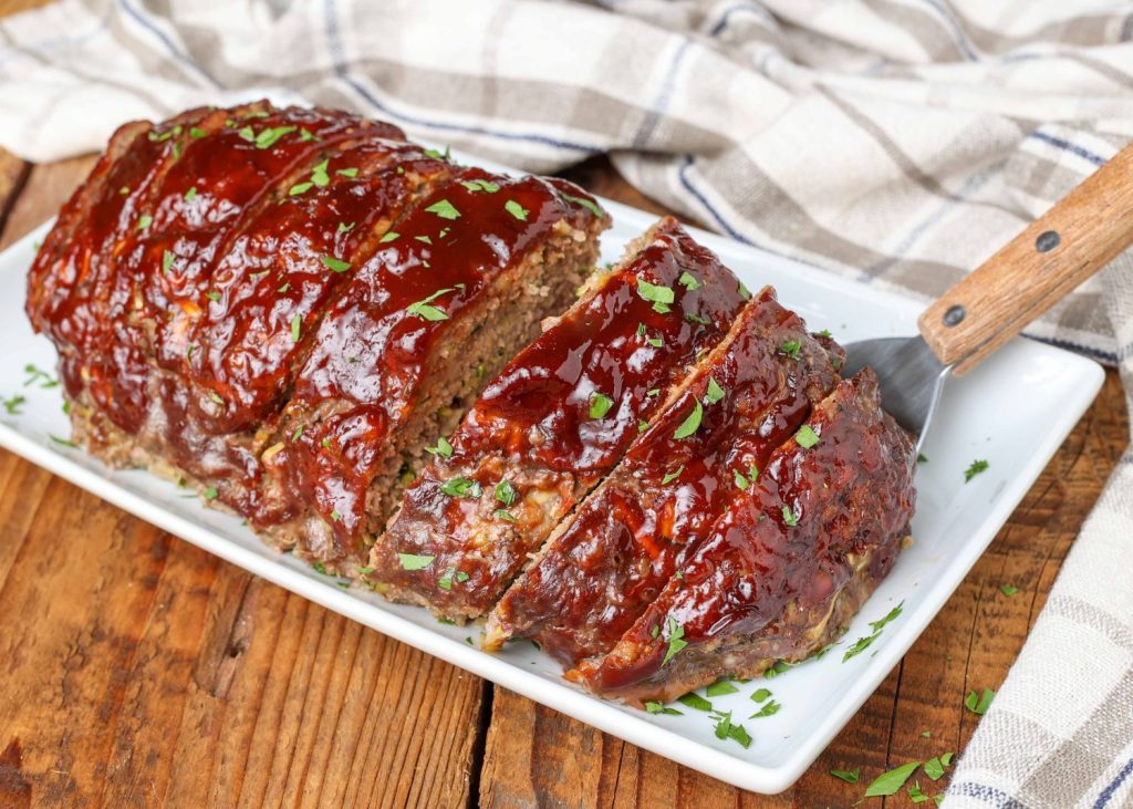 Overhead shot of large sliced meatloaf containing zucchini, topped with BBQ sauce, served on a long white rectangular tray with a striped white and gray hand towel