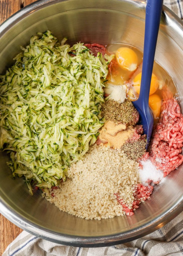 Overhead process shot of meatloaf ingredients (ground beef, shredded zucchini, eggs, miscellaneous spices) in a steel bowl with a blue spatula