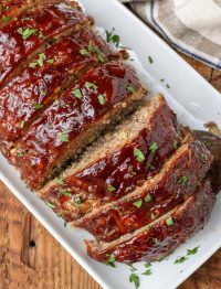 Overhead diagonal shot of large sliced meatloaf containing zucchini, topped with BBQ sauce, served on a long white rectangular platter