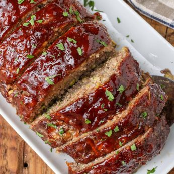 Overhead diagonal shot of large sliced meatloaf containing zucchini, topped with BBQ sauce, served on a long white rectangular platter