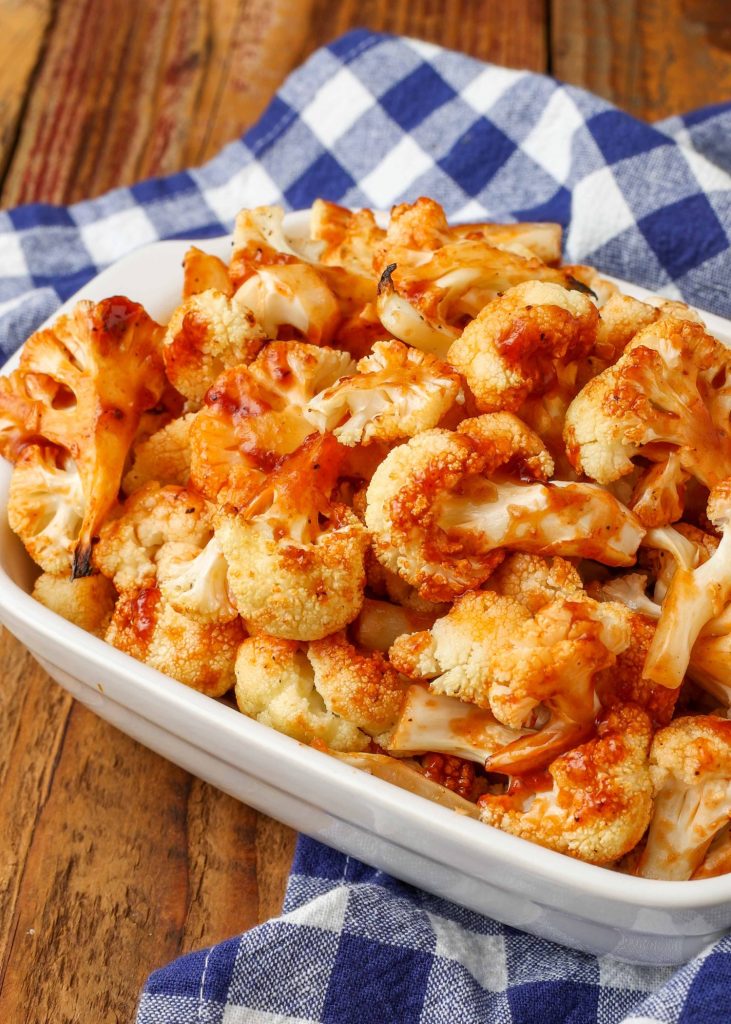 BBQ cauliflower is piled high in a white ceramic baking dish with a blue and white checkered tea towel visible in the background