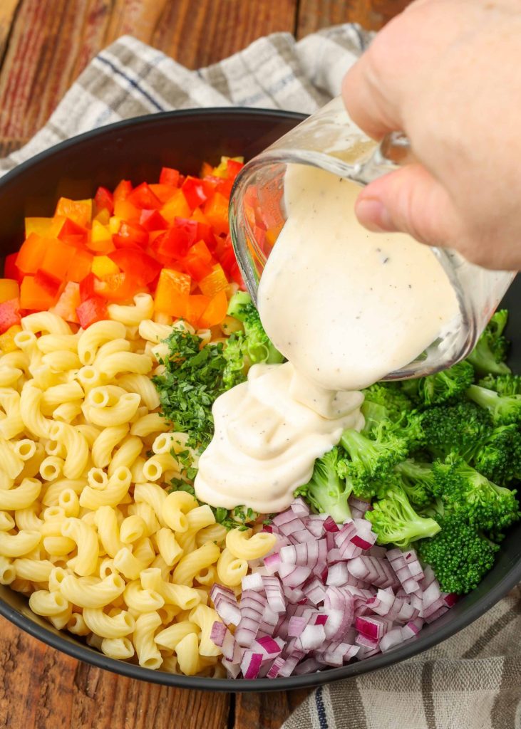 Pasta, chopped bell peppers, broccoli florets, and diced onions, portioned in a large black bowl; creamy salad dressing is being poured over the bowl