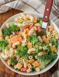 Overhead vertical shot of broccoli pasta salad, served in a white bowl with a silver spoon and a checkered white and gray towel