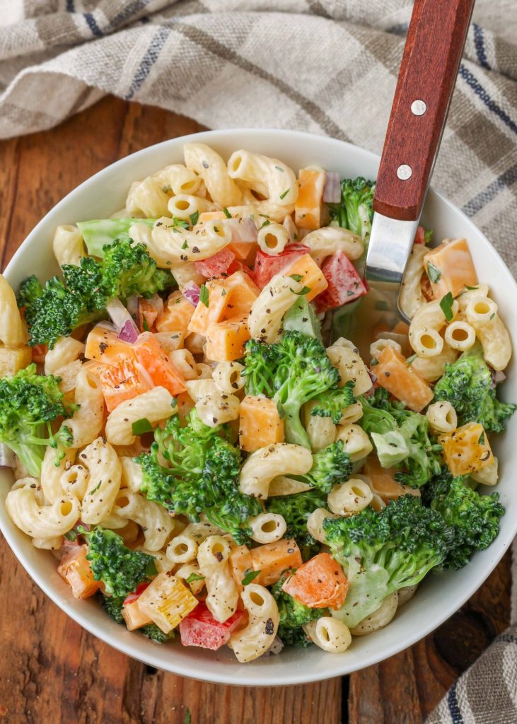 Overhead vertical shot of broccoli pasta salad, served in a white bowl with a silver spoon and a checkered white and gray towel