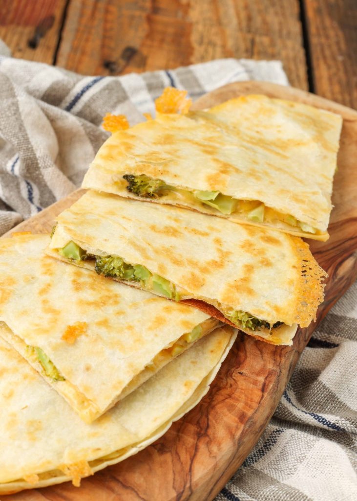 Overhead shot of quartered cheesy broccoli quesadillas stacked on top of one another, served on a cutting board