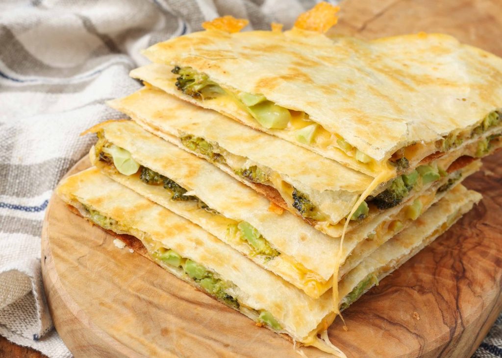 Overhead shot of quartered cheesy broccoli quesadillas stacked on top of one another, served on a cutting board