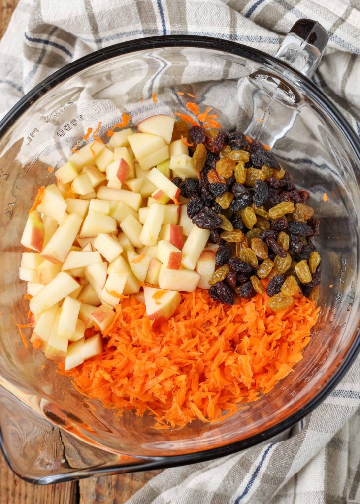 Overhead shot of chopped apples, shredded carrots, and mixed raisins in a glass bowl