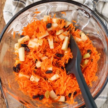 Overhead shot of tossed carrots, apples, and mixed raisins, served in a glass bowl