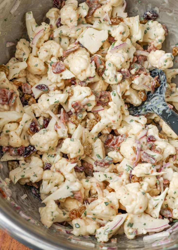 a metal mixing bowl contains the ingredients for the salad, freshly mixed with dressing. visible are bits of bacon, cauliflower florets, and craisins.