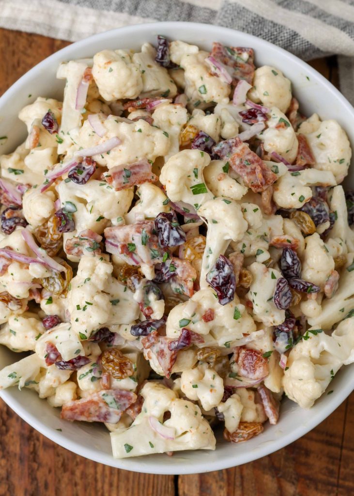 A top down shot of a serving of cauliflower bacon salad in a white bowl with a grey and white striped napkin visible in the background.