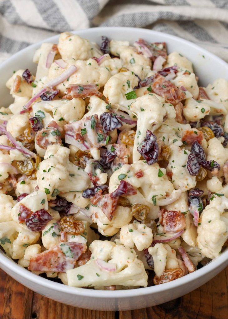 A close up shot of a serving of cauliflower bacon salad in a white bowl with a grey and white striped napkin visible in the background.