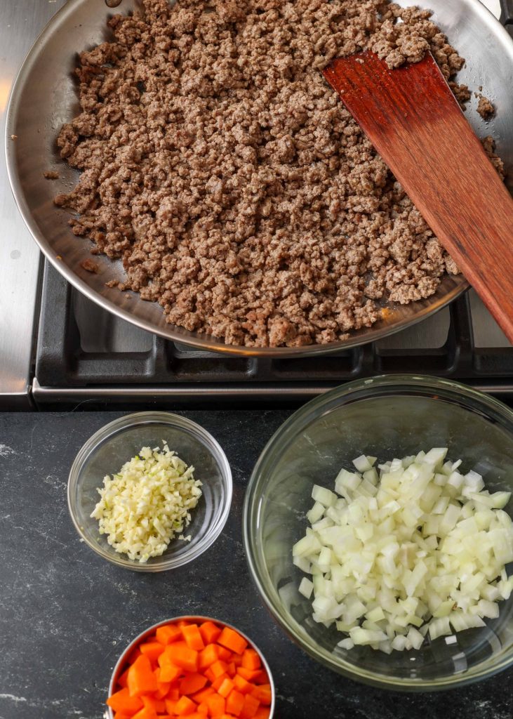 The cooked and crumbled ground beef is beside three small glass bowls with the fresh ingredients for the filling.
