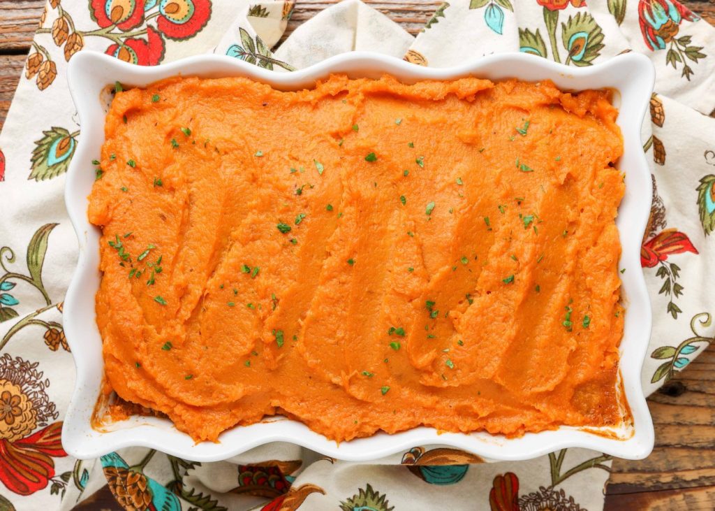 A horizontally aligned photo of a white baking dish, loaded with ground beef casserole topped with mashed sweet potatoes.
