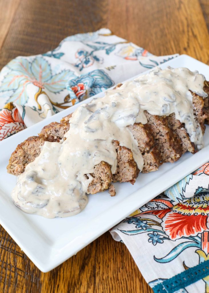A vertically aligned photo of a sliced meatloaf with white gravy with mushrooms in it, on a long white serving platter over a wooden tabletop.