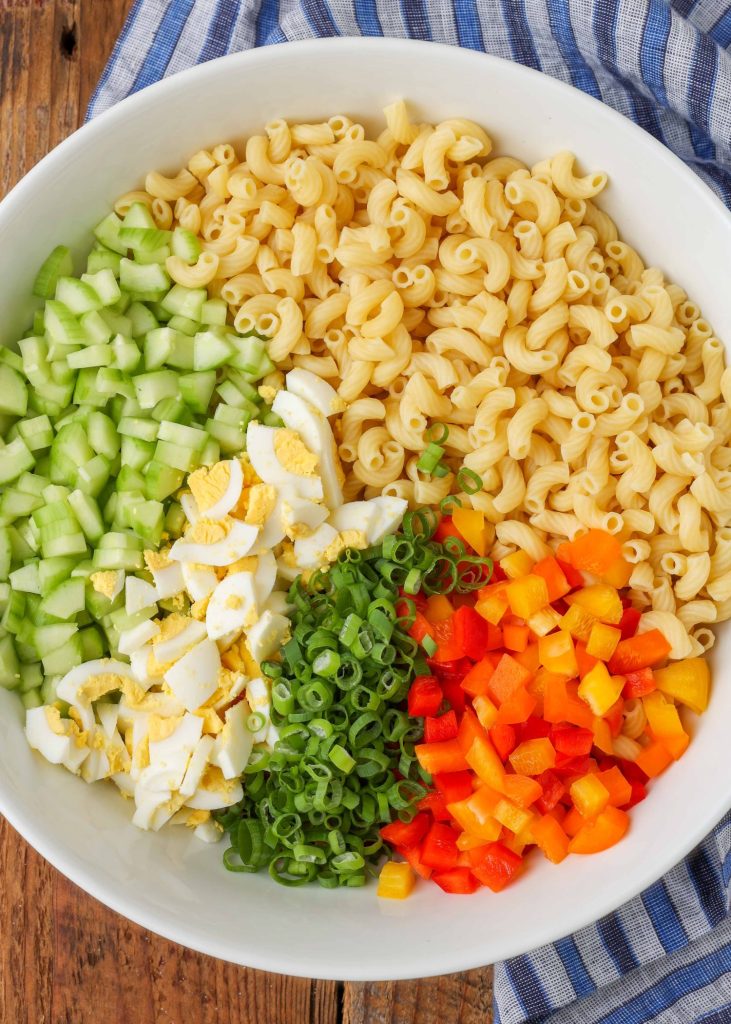 All of the ingredients for macaroni salad have been artfully arranged in a white bowl, showing the colorful vegetables and tender pasta.