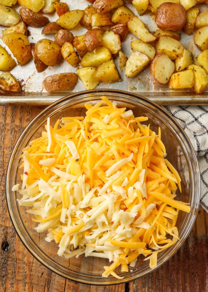 A bowl of shredded cheeses stands beside a metal sheet pan with roasted potatoes.