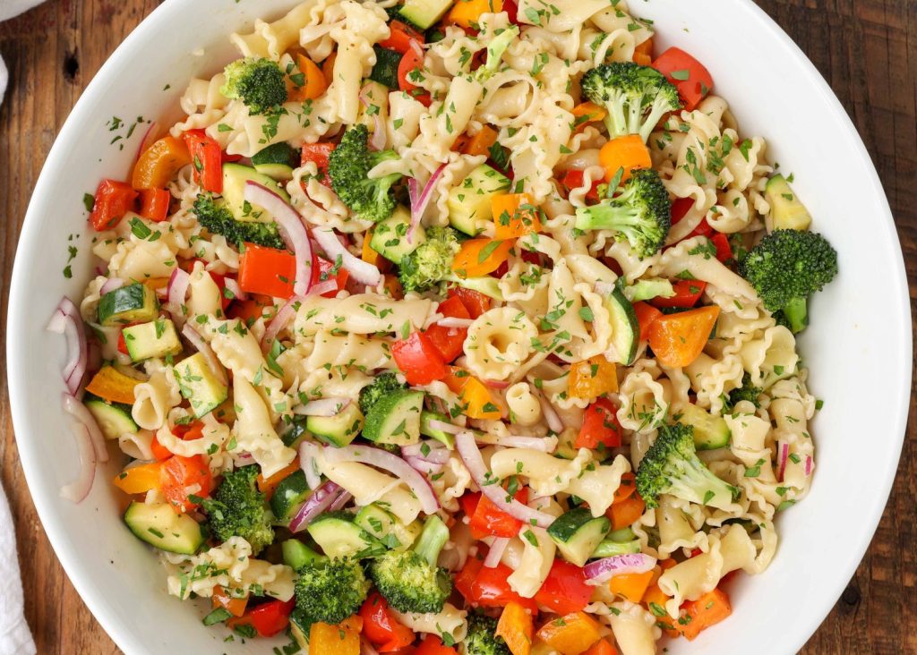 a horizontally aligned photo of a bowl of salad with broccoli, peppers, noodles, red onions, and zucchini glistens with the zesty oil and vinegar dressing.