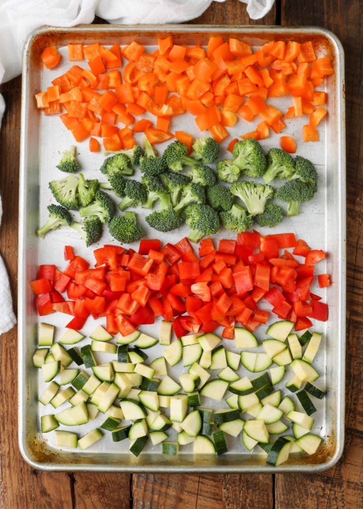 Orange bell peppers, green broccoli florets, red bell peppers, and sliced zucchini have been arranged in rows on a metal sheet pan.