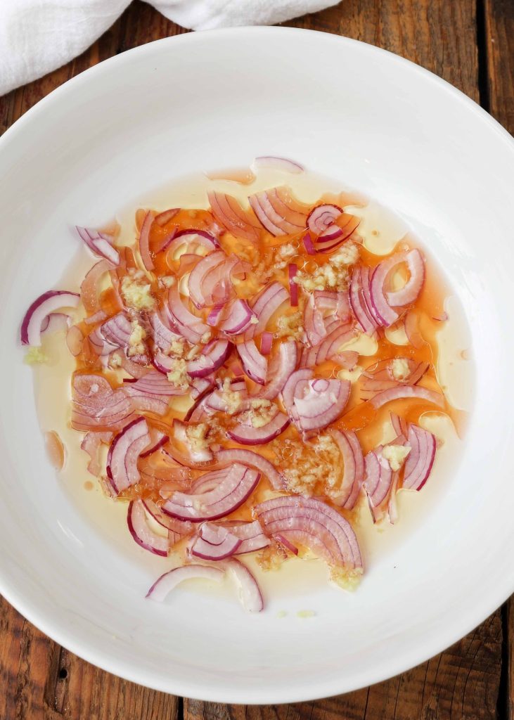 red onions and vinegar, oil, and garlic have been added to a white mixing bowl to create the dressing for this salad.