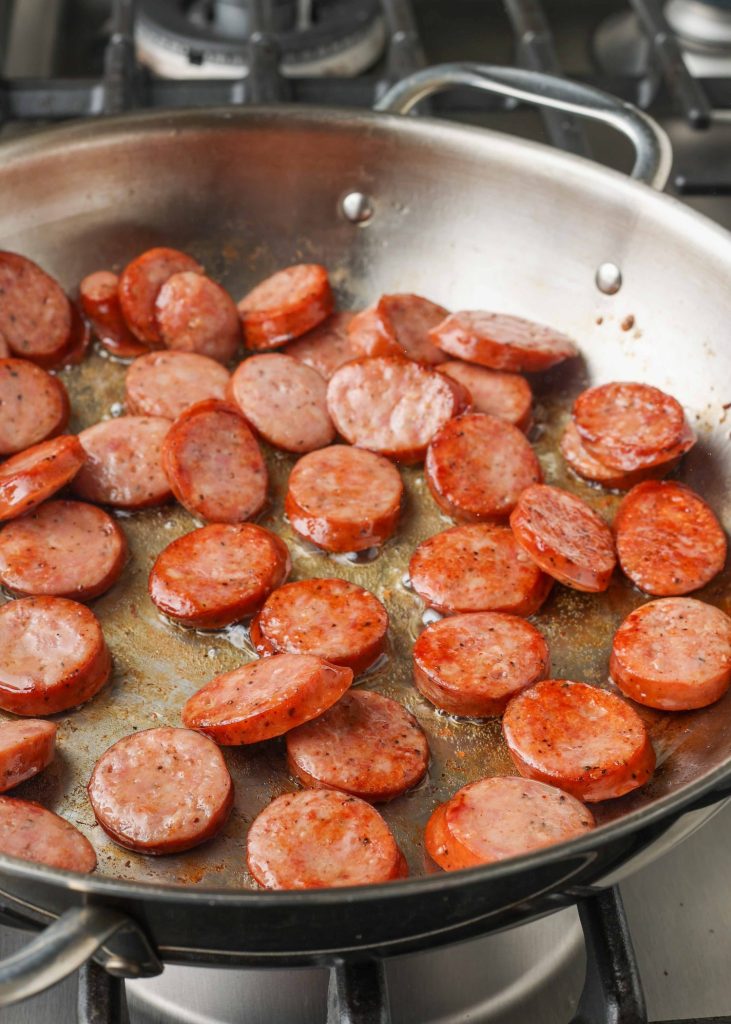 Slices of kielbasa sausage sizzle in a metal pan in this vertically aligned photo.
