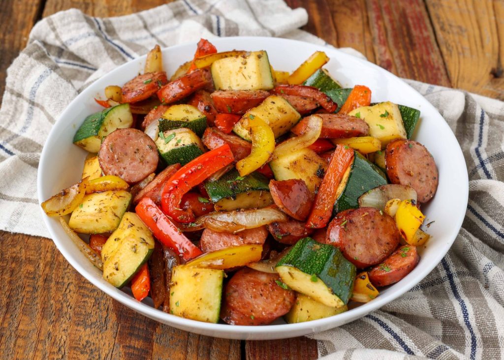 a horizontally aligned photo of a white bowl on a wooden tabletop, full of sausage, zucchini, peppers, and onions.