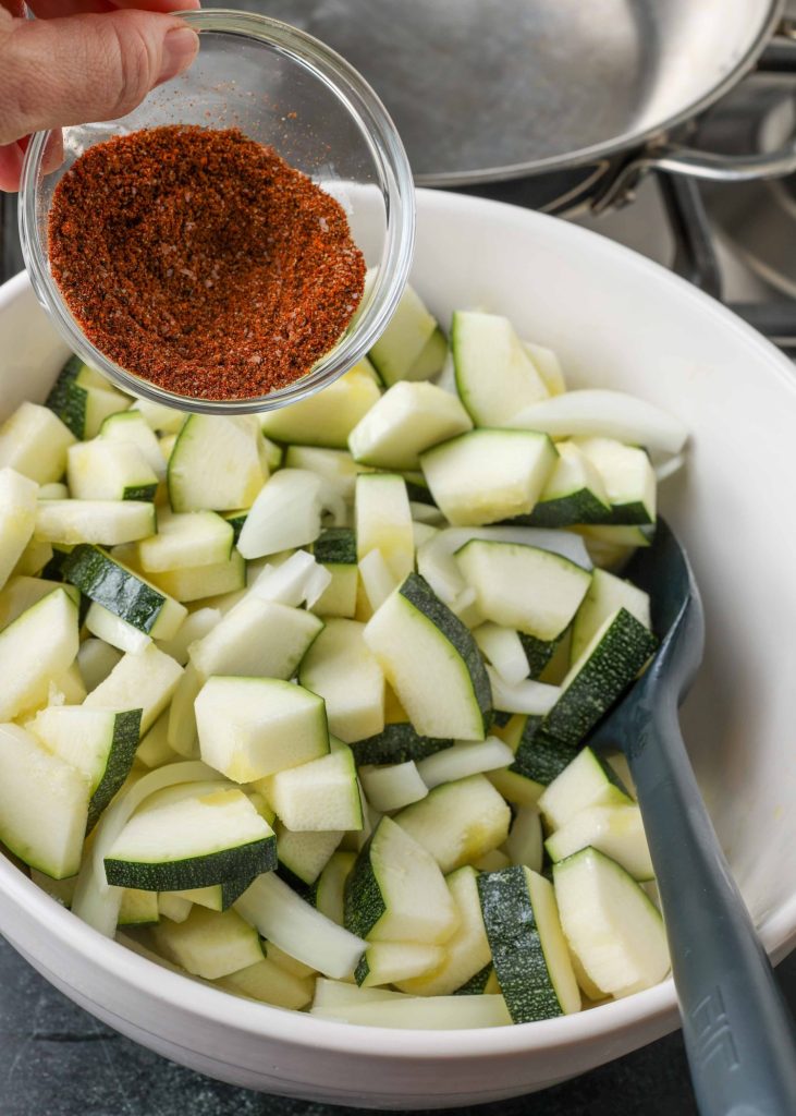 chopped zucchini pieces are in a white bowl, with a smaller glass bowl of spices about to be poured over them.